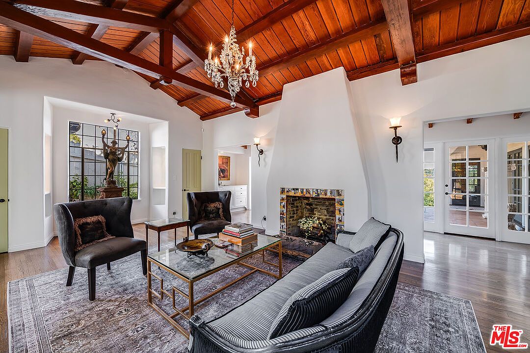 The great room at 1046 Chautauqua. A cathedral wood-beamed ceiling rises over a stone fireplace, leaded-glass windows, dark wood floors, and a curved velvet sofa beneath a brass-and-crystal chandelier.